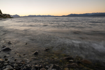 Long exposure shot of Nahuel Huapi lake at sunset. Beautiful blurred water effect, the rocky shore and Andes mountains in the horizon with a magical dusk light.