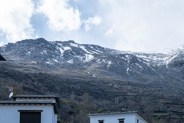 Bottom-up view to rocky mountains partly covered by snow with cloudy sky in background and part of white buildings in the foreground