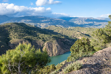 Fototapeta na wymiar Breath-taking view from the top on green lake surrounded by picturesque mountain alley with trees and bushes in sunny summer day with blue sky and white clouds in background.