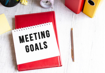 Notepad with the text MEETING GOALS on a wooden table. Red diary and pen.
