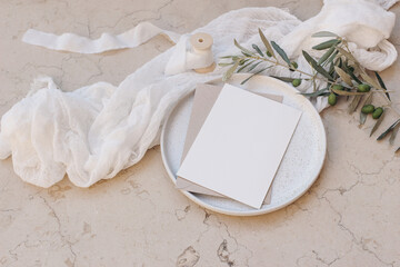 Mediterranean wedding stationery. Greeting card, invitation mock up. Envelope on plate.Beige marble background. Olive tree branches, silk ribbons. White muslin table runner. Summer design. Top view.