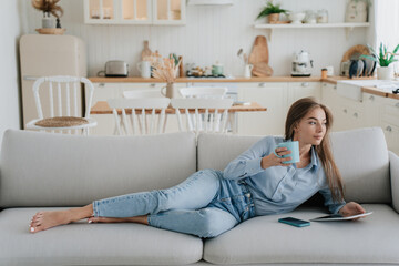Beautiful blonde european young woman with long hair, lying on the sofa in a blue shirt and jeans in the kitchen with a tablet and a phone, holding a coffee mug in her hand and looking away calmly.