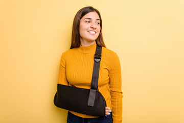 Young caucasian woman with broke arm isolated on yellow background looks aside smiling, cheerful...