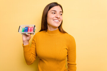 Young caucasian woman holding a batteries box isolated on yellow background looks aside smiling, cheerful and pleasant.