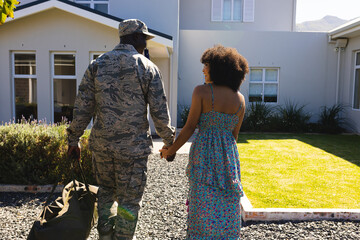 Rear view of multiracial army soldier in camouflage holding wife's hand and walking towards house