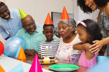 Senior woman wearing party hat blowing candles while celebrating birthday with multiracial family