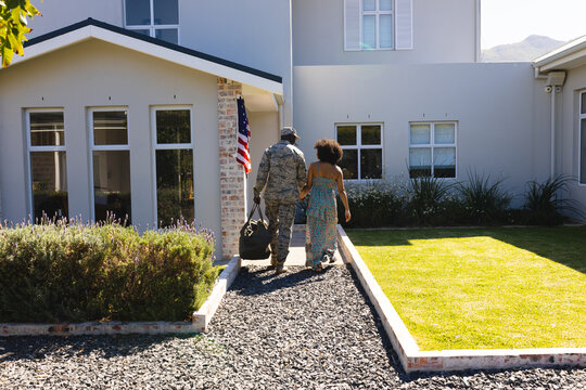 Rear view of multiracial army soldier in camouflage holding wife's hand and going inside house