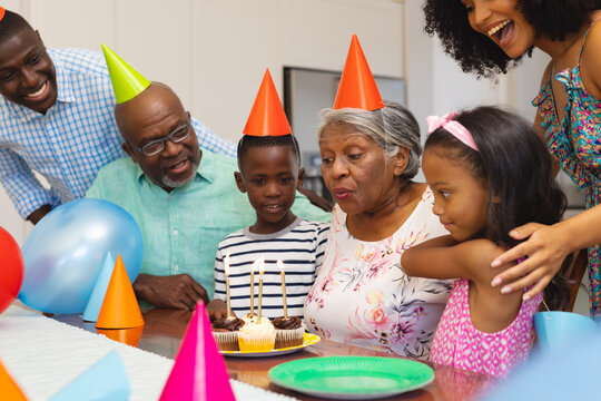 Senior woman wearing party hat blowing candles while celebrating birthday with multiracial family - Powered by Adobe