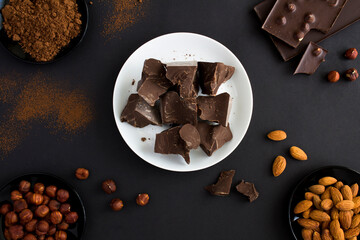 Top view of chunks of dark chocolate on white plate, chocolate, cocoa powder and nuts on the black background. Chocolate confectionery background.