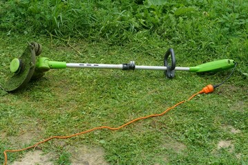 dirty modern handy hand trimmer with electric orange cord turned on lies on mowed green grass...