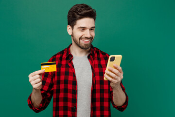 Young happy smiling man he 20s wearing red shirt grey t-shirt using mobile cell phone hold credit bank card doing online shopping order delivery booking tour isolated on plain dark green background.