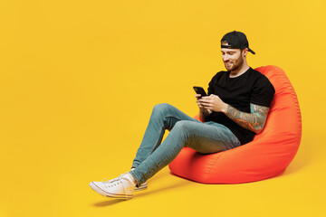 Full body young bearded tattooed man 20s he wears casual black t-shirt cap sit in bag chair hold in hand use mobile cell phone isolated on plain yellow wall background studio People lifestyle concept.