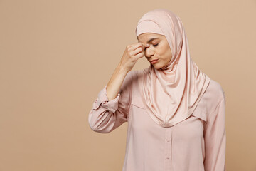 Young arabian asian muslim woman she wear abaya hijab pink clothes keep eyes closed rub put hand on nose isolated on plain pastel beige background. People uae middle eastern islam religious concept.