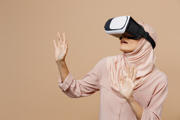 Young arabian asian muslim woman she wear abaya hijab pink clothes watching in vr headset pc gadget isolated on plain pastel light beige background. People uae middle eastern islam religious concept.