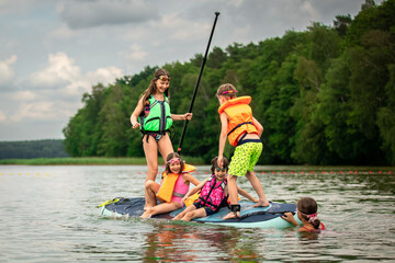Children having fun on SUP board waving it in water of lake and laughing on cloudy summer day...