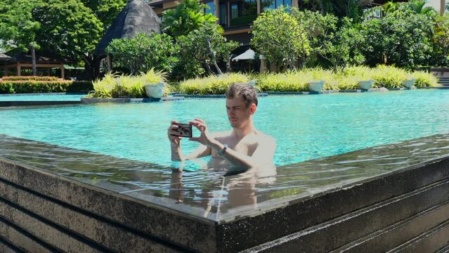 Young serious man take pictures, make photo on smartphone standing in swimming pool at luxury tropical hotel, resort. Influencer, blogger create summer vacation or holiday content for social media.
