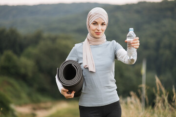 Front view of charming woman in hijab and sport clothes standing at green park and holding yoga mat...