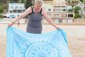 Short haired woman with glasses and wearing printed dress spreads blue and white towel on the beach.