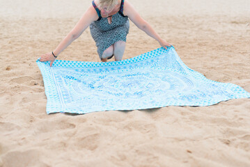 Knee level, medium long shot image of a woman on the beach wearing a geometric patterned dress stretching out a blue towel with geometric shapes for sunbathing.sunbathe