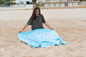 Knee-level, long shot of a long-haired boy laying out his towel on the beach to sunbathe.