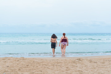 Boy with long hair and bermuda shorts and a young adult entering the sea for a swim. There is copy space.