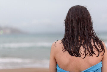Teenager with long hair looking towards the horizon on the beach after bathing, while drying himself wrapped in a blue towel.