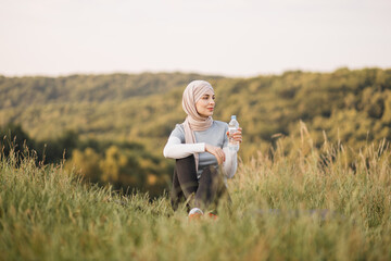 Young sporty muslim woman drinking water from plastic bottle, taking rest break after working out, exercising, sitting on mat on green grass in park outdoors. Water balance. Horizontal shot.