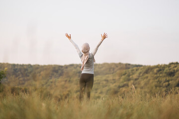 Fototapeta premium Arabian young woman is practicing yoga in the summer park. A Muslim woman is looking aside exercising outdoor and breathing fresh air outstretching arms . Fit arabic woman training in a park