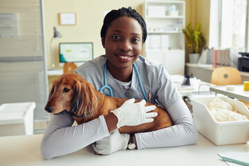 Portrait of smiling young veterinarian hugging dog and looking at camera in vet clinic