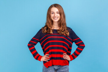Portrait of smiling positive woman wearing striped casual style sweater looking at camera, keeping hands on hips, having confident expression. Indoor studio shot isolated on blue background.