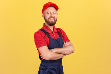 Confident smiling handyman wearing overalls standing with crossed arms, looking at camera,...