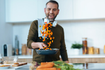 Handsome mature man sautéing vegetables in the pan while having fun in the kitchen at home.