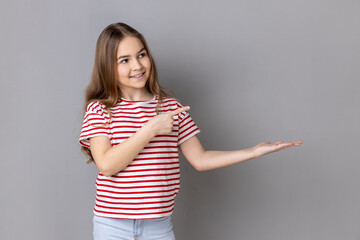 Portrait of little girl wearing striped T-shirt presenting advertising area on her palm and pointing to copy space, holding empty place for commercial. Indoor studio shot isolated on gray background.