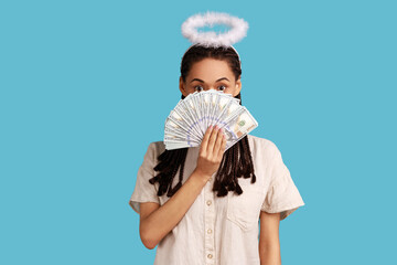 Angelic happy rich woman with black dreadlocks and nimbus on head peeking out of dollar banknotes,...
