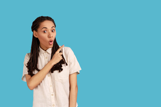 Surprised woman with black dreadlocks keeps mouth opened pointing finger away on blank space, shows amazing offer, wearing white shirt. Indoor studio shot isolated on blue background.