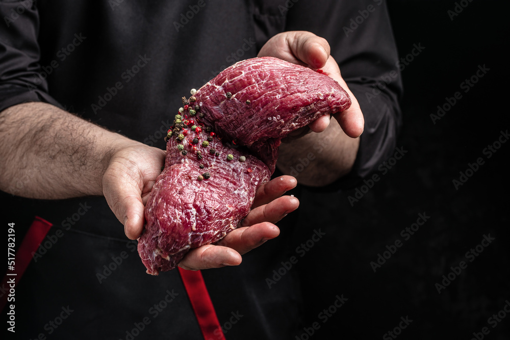 Wall mural chef salts steak in a freeze motion with rosemary and spices. preparing fresh beef or pork on a dark