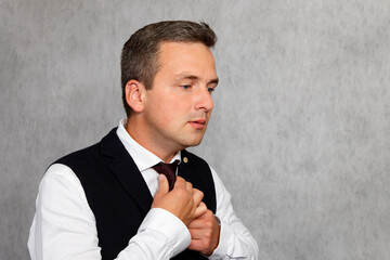 A handsome man in a white shirt and dark vest ties a tie.