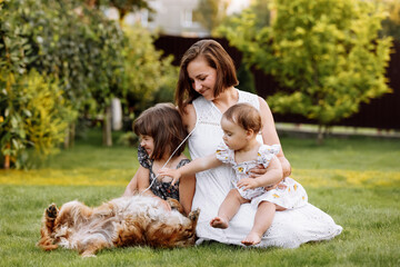 Family day, mother's day. Beautiful smiling young mom and two child daughters cuddling happy domestic dog on the backyard lawn. Idyllic family having fun with pet outdoors on summer holiday.