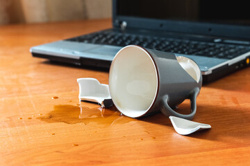 Broken cup and spilled tea or coffee near laptop in the office