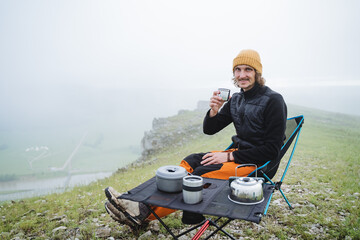 A happy young man on a hike in the mountains sits at a table for dinner, a man holds in his hand a mug of hot tea, delicious food in a campsite in nature, camping utensils.