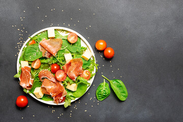 Fresh juicy green salad with cherry tomatoes, goat cheese and prosciutto on black stone background top view. Healthy tasty food concept.