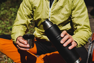 Thermos with tea, a person holds a bottle of water, a black metal thermos with a hot drink, breakfast in nature, tourist dishes, pour tea.