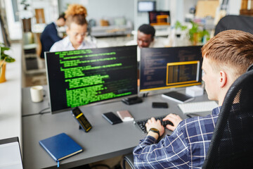Rear view of young IT specialist writing codes of software sitting at office desk in front of computer monitors