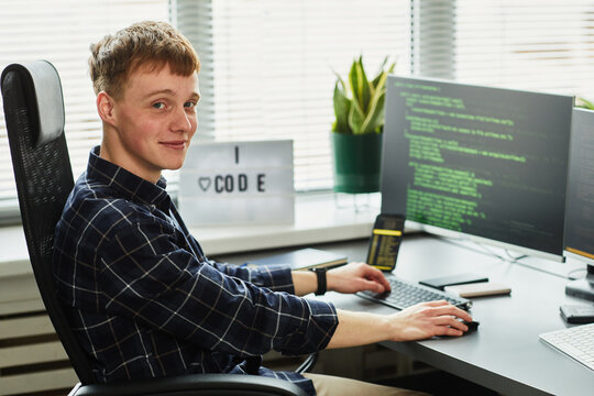 Portrait of young programmer looking at camera while sitting at his workplace with computer monitor and writing codes