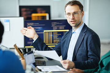 Mature developer pointing at computer monitor with interface and giving report to his colleagues at...