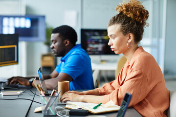 Obraz premium Young woman typing on laptop working with new interface while sitting at table at office with her colleague in background