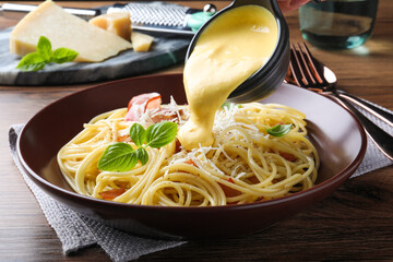 Pouring tasty cheese sauce onto spaghetti with meat on wooden table, closeup