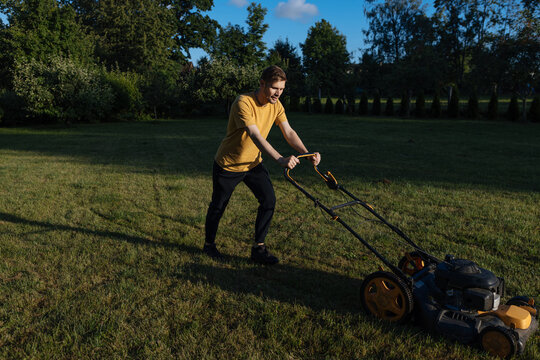 A Man Mows Grass In The Yard Of A House. A Young Man Pushes A Lawn Mower. Difficulty Cleaning The Yard. An Exhausted Man Doing Home Work. Summer Fatigue. Shoot From Above.