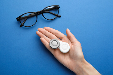 Woman holding case with contact lenses near glasses on blue background, top view