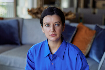 Portrait of a beautiful young woman sitting on couch at home. Caucasian girl with blue sad eyes in...